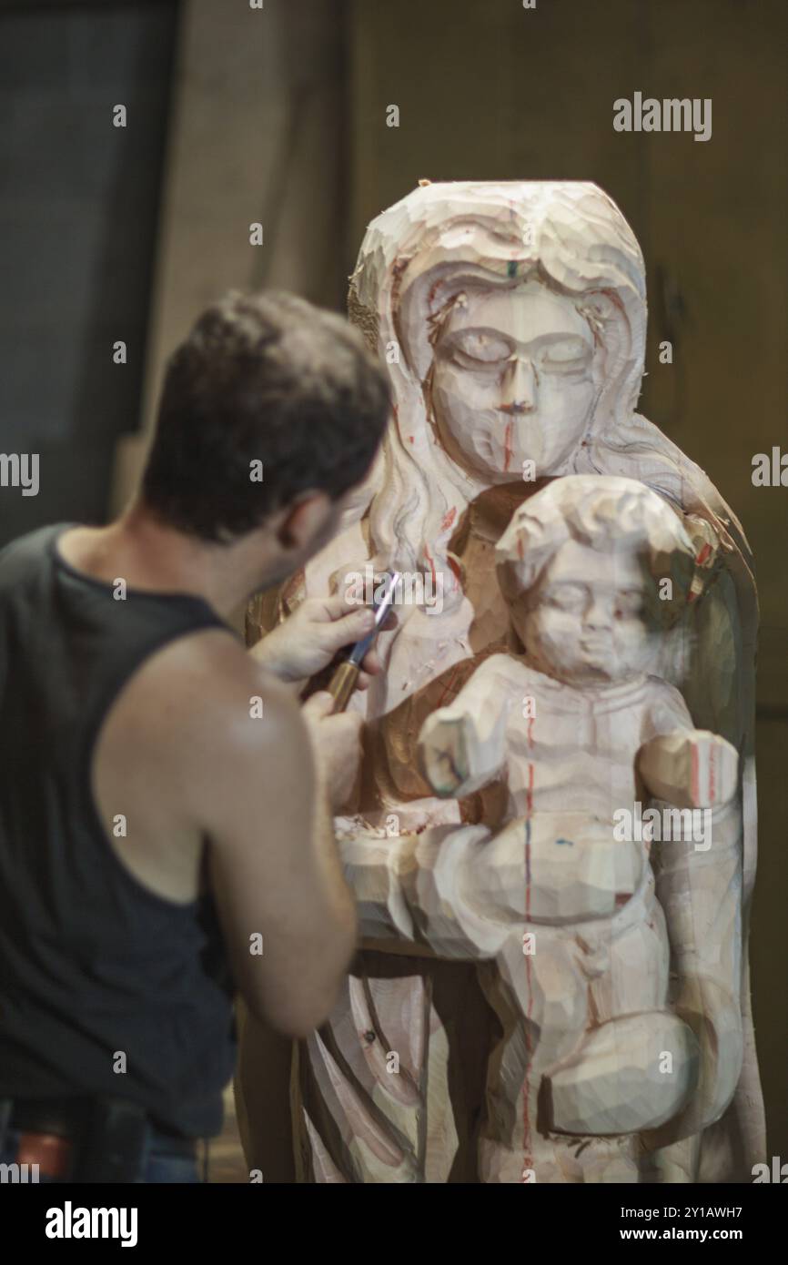 Ein Handwerker schnitzt akribisch eine Holzskulptur einer Frau, die in einer Werkstatt ein Kind hält Stockfoto