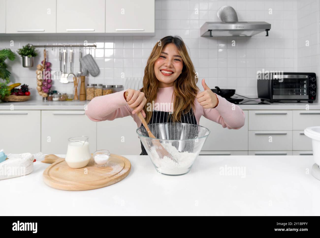 Eine glückliche Frau in einer Schürze, die Teig in einer klaren Glasschale mit Spachtel mischt. Sitzen Sie in einer sauberen und modernen Küche, lächeln Sie, während Sie den Daumen nach oben heben. Stockfoto