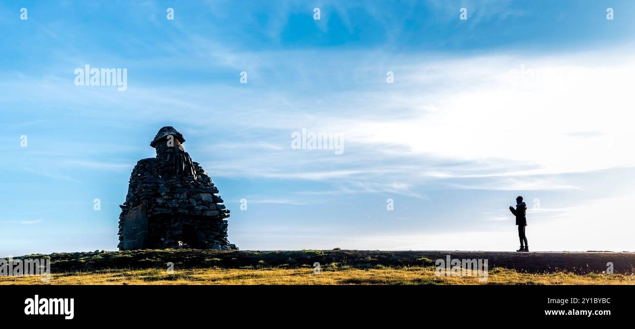 Ein Mann steht auf einem Hügel und blickt auf eine große Steinstruktur. Bardur Snaefellsas Statue. Der Himmel ist blau und die Sonne scheint hell. Konzept von Stockfoto