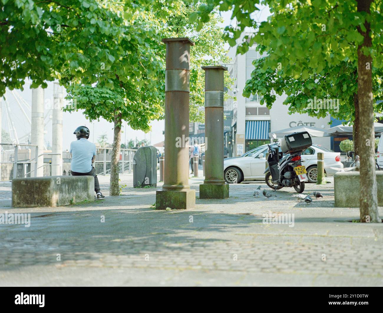 Ein Lieferfahrer wartet an einem heißen Tag im Schatten seines Mopeds in Cardiff Bay, während Tauben in der Nähe füttern. Stockfoto