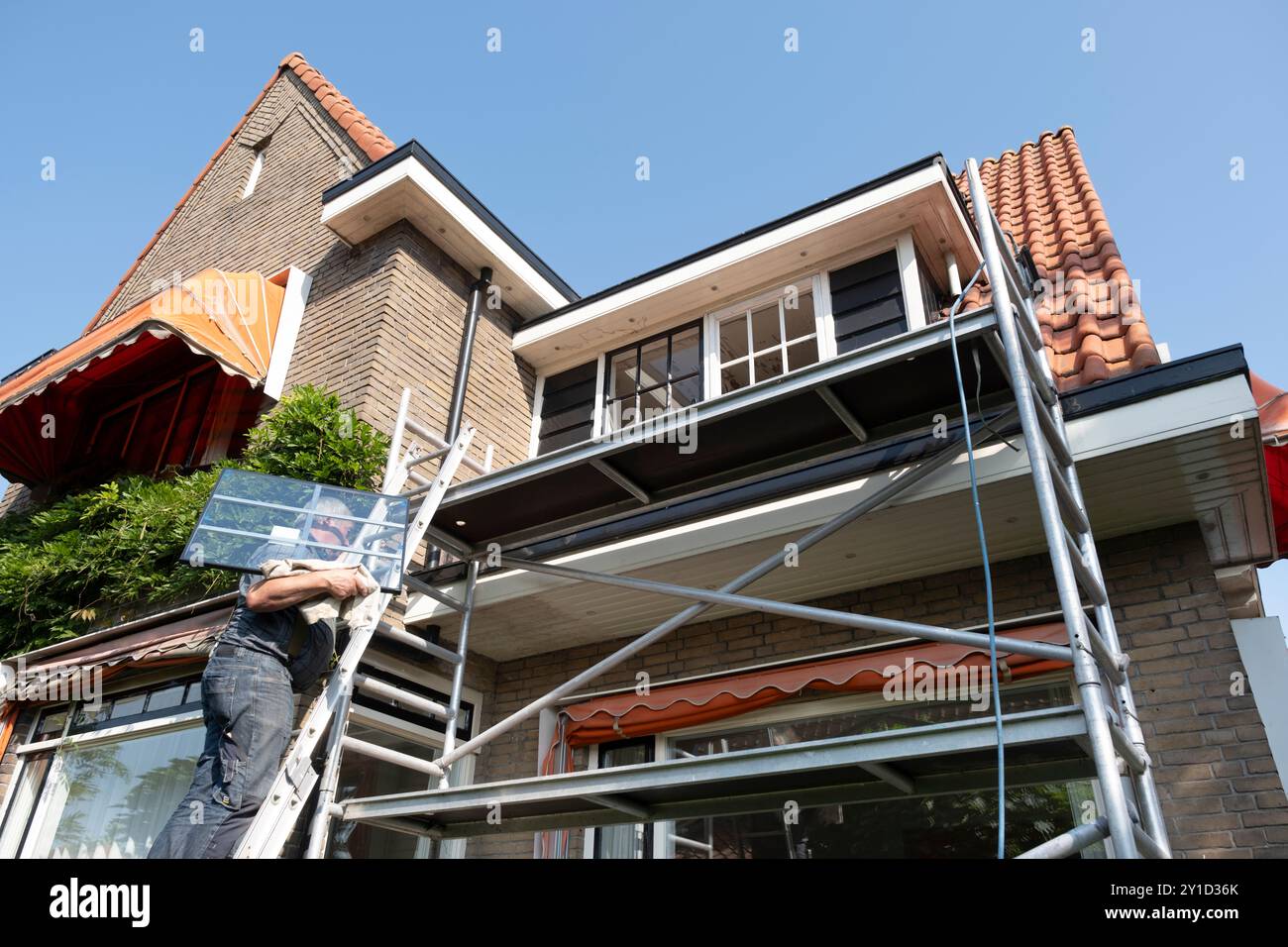 Der Bauarbeiter steigt eine Leiter mit einem neuen Fenster mit Stangenteilung hinauf. Renoviertes Haus mit Doppelverglasung Stockfoto