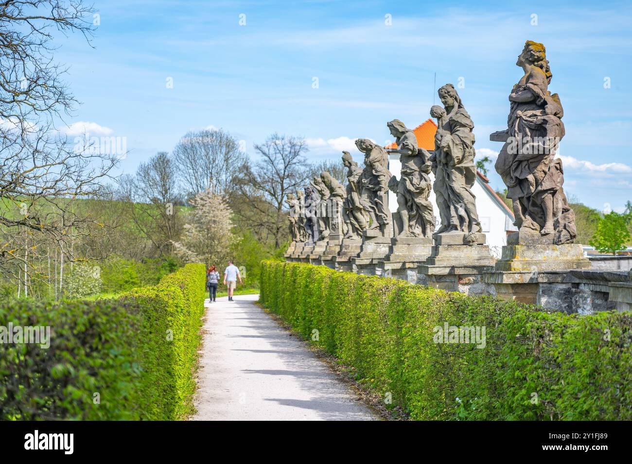 Besucher schlendern auf einem mit barocken Statuen geschmückten Weg im Krankenhauskomplex von Kuks, der sich vor einem klaren blauen Himmel und blühendem Grün befindet und historische Kunst und Architektur zeigt. Stockfoto