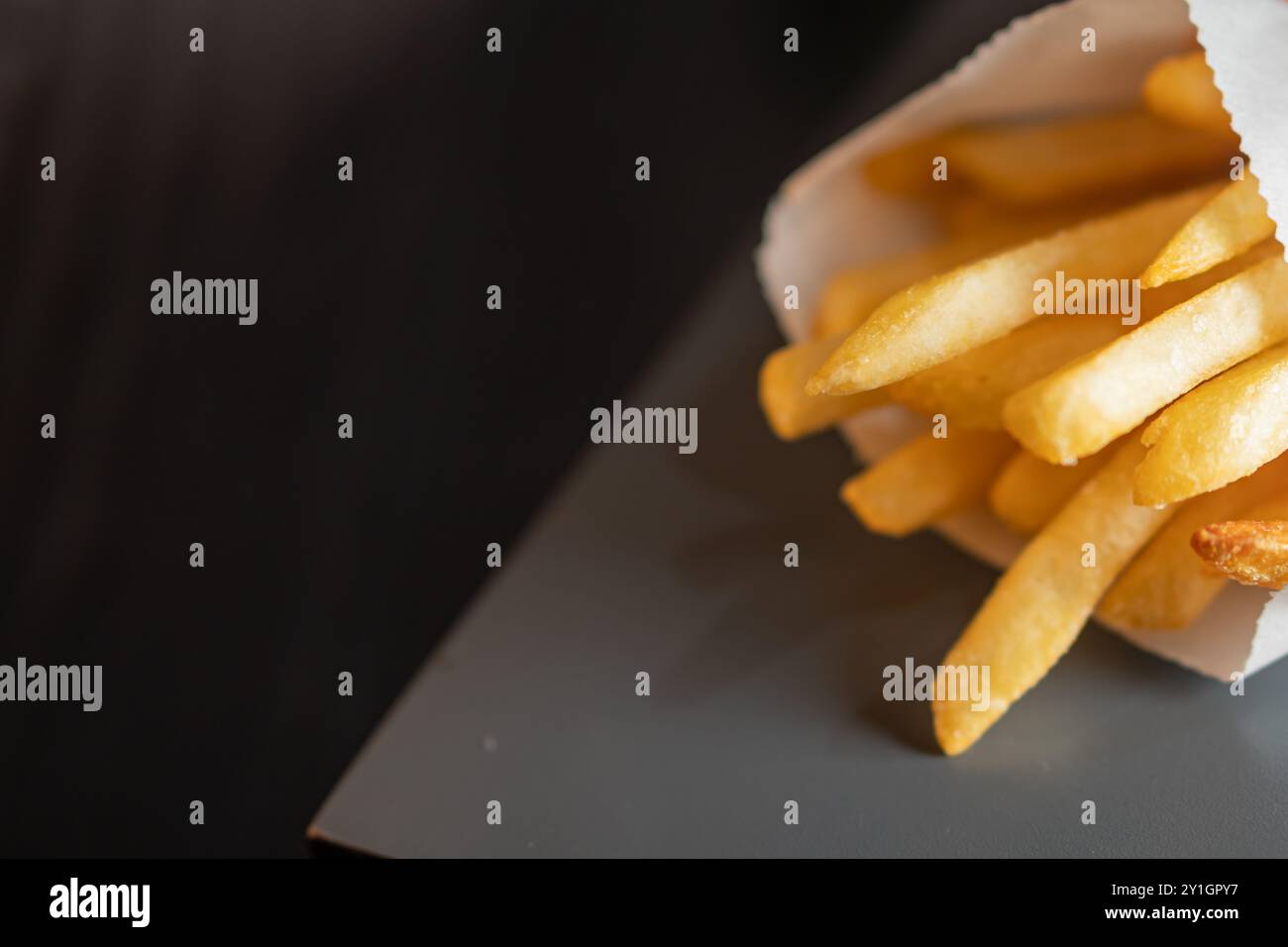 Eine Nahaufnahme einer Portion goldener Pommes frites in einer Papiertüte, die auf eine dunkle Oberfläche gelegt wurde. Die Pommes frites sind knusprig und frisch gekocht, mit einem weichen Fokus Stockfoto