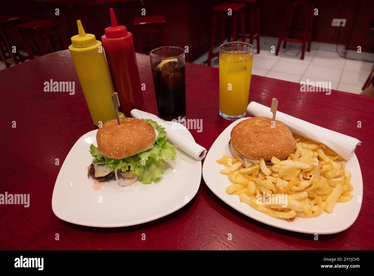 Mahlzeit für zwei in einem Fast Food: Zwei leckere gegrillte Burger mit Pommes frites, Limonade und Orangensaft auf weißen Gerichten und rotem Holztisch, mit Plastikscheiben Stockfoto