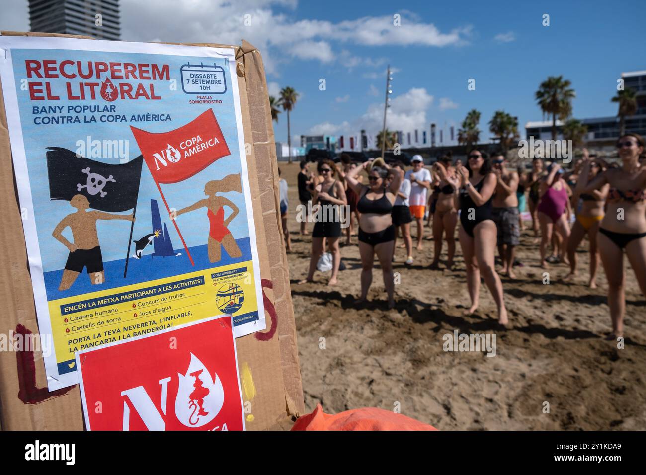Das Poster, das für die Segelveranstaltung des America's Cup aufruft, ist zu sehen, wenn die Demonstranten an einem improvisierten Tanzkurs am Strand teilnehmen. Hundert Aktivisten und Einwohner Barcelonetas haben am Strand von Somorrostro in Barceloneta eine Menschenkette gebildet, um gegen die Segelveranstaltung des America's Cup zu protestieren. Nach Aussagen der Demonstranten untergräbt die Veranstaltung dieses großen Sportereignisses die Integrität der Arbeiterviertel, ignoriert Forderungen nach sofortigen Maßnahmen zur Verringerung der Tourismuswirtschaft und enteignet den Bürgern massiv den öffentlichen Raum. All dies ohne wirtschaftlichen Nutzen Stockfoto