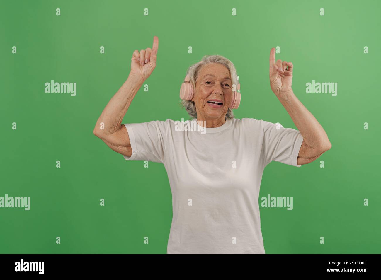 Eine freudige Seniorin tanzt glücklich mit Ohrenschützern auf einem hellgrünen Hintergrund Stockfoto