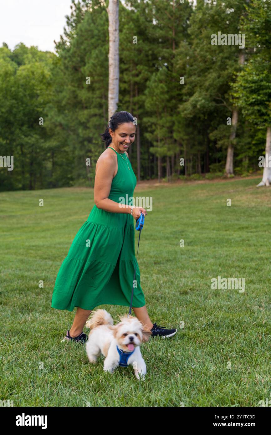 Eine Frau in einem grünen Kleid geht mit ihrem Hund in einem Park. Der Hund trägt ein blaues Gurtzeug Stockfoto