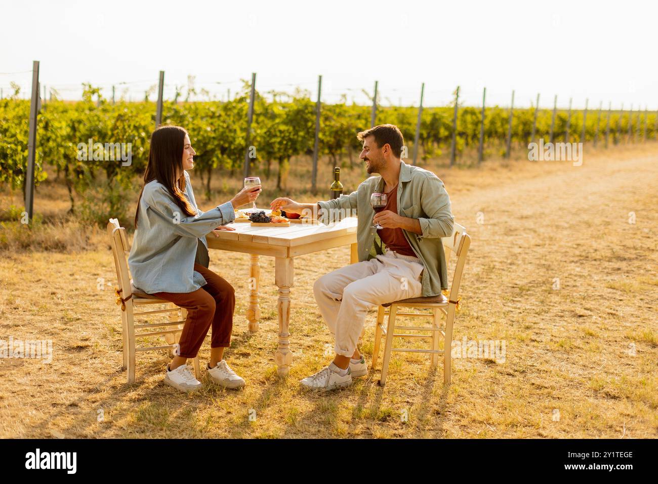 Ein paar entspannen sich an einem rustikalen Tisch in einem sonnigen Weinberg, genießen Wein und Gourmet-Snacks. Sie teilen Lachen und Freude, umgeben von üppigen Weinstöcken und dem Stockfoto