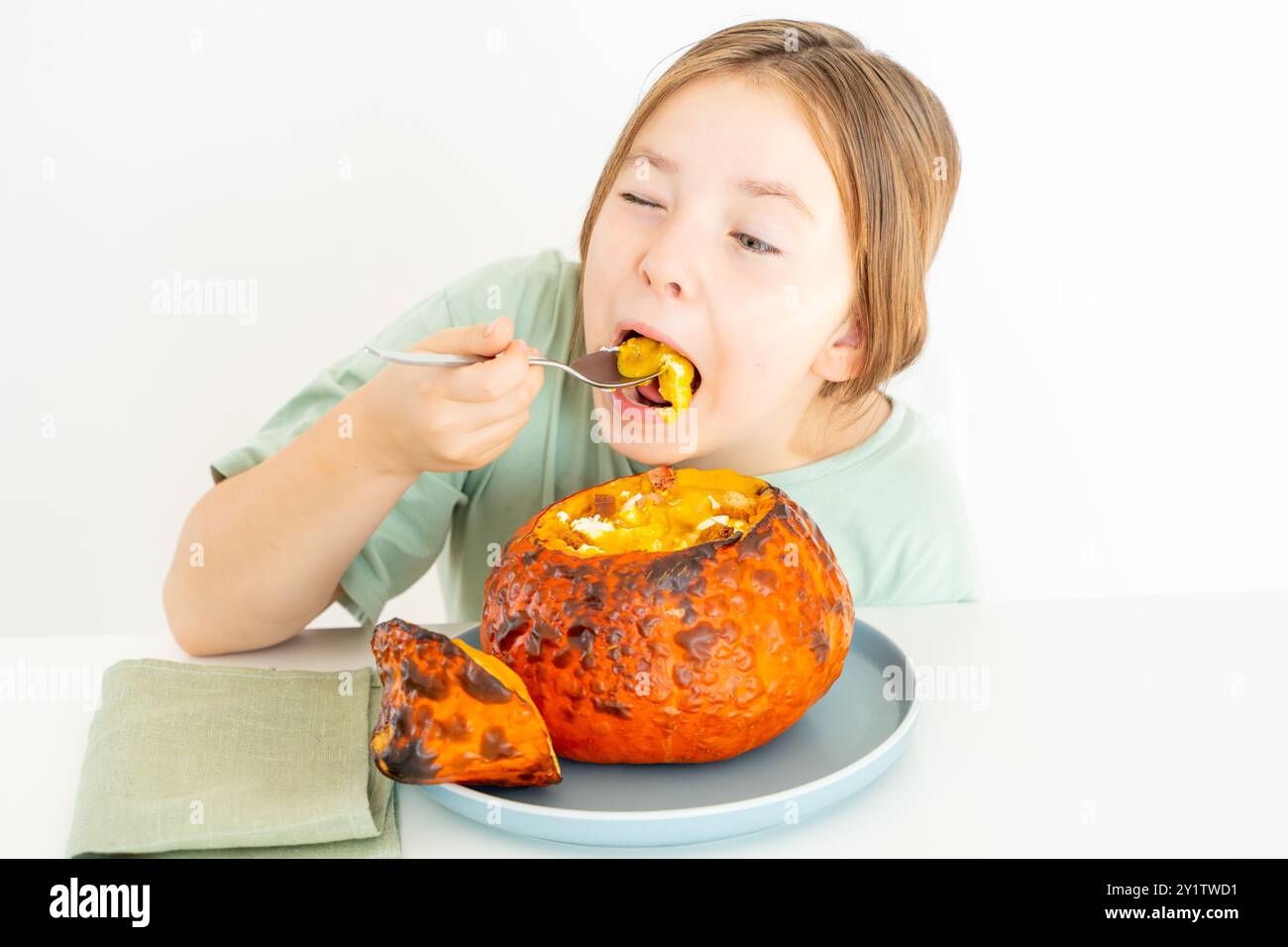 Kleines Mädchen im Teenageralter isst Kürbissuppe, serviert in gebackenem Kürbis. Mädchen in grünem T-Shirt und Herbstschale auf weißem Hintergrund. Hochwertige Pho Stockfoto