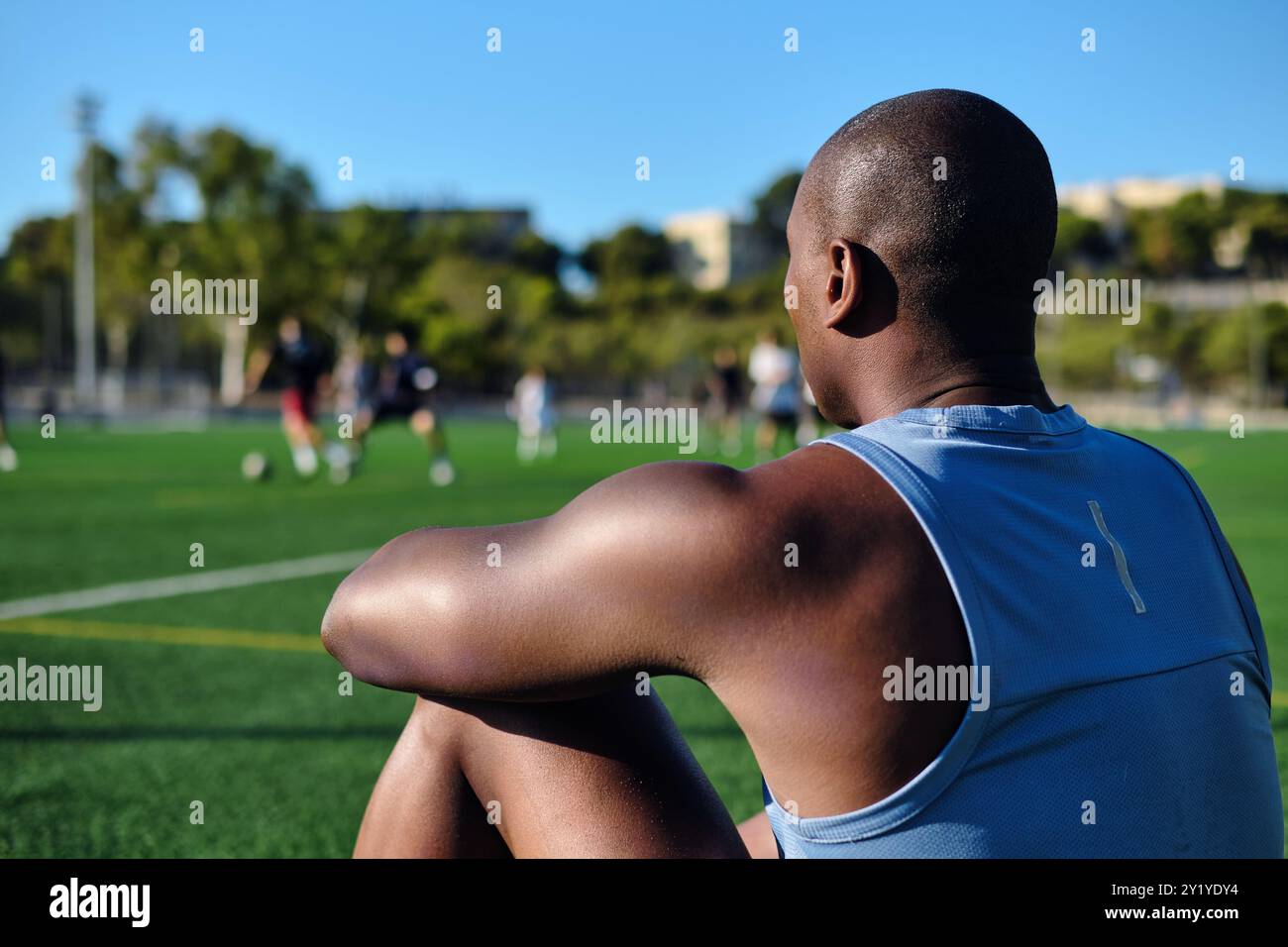 Junger Fußballspieler tragen ein blaues, athletisches Tanktop, das auf einem grasbewachsenen Sportfußballfeld sitzt und sich nach dem Spiel oder Workout ausruht. Pause, Sport Stockfoto