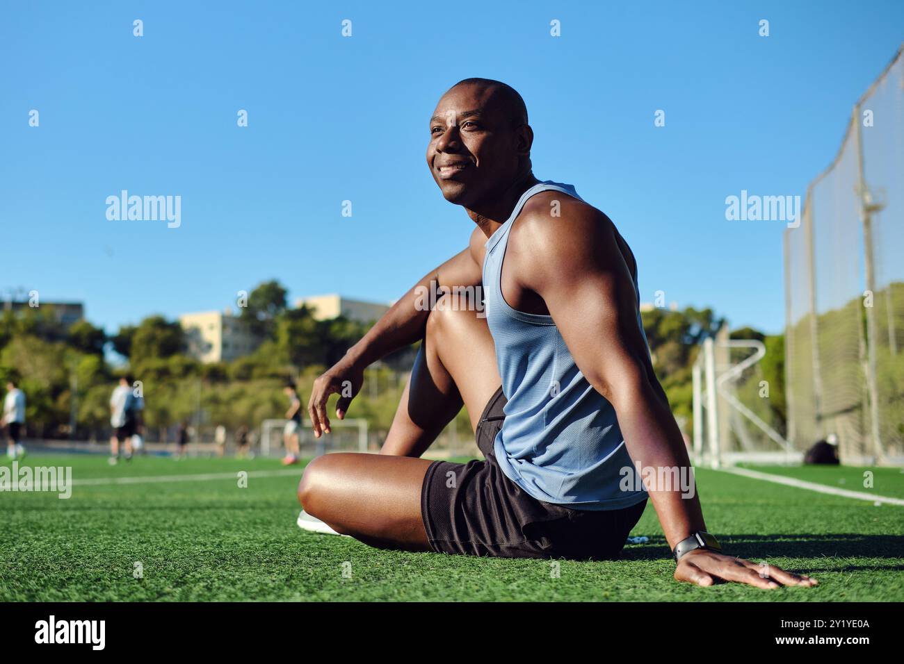 Sportlicher Mann Fußballspieler tragen blaues, sportliches Tanktop und schwarze Shorts, sitzen auf einem grasbewachsenen Fußballfeld, ruhen sich nach dem Spiel oder Workout aus, sehen aus Stockfoto