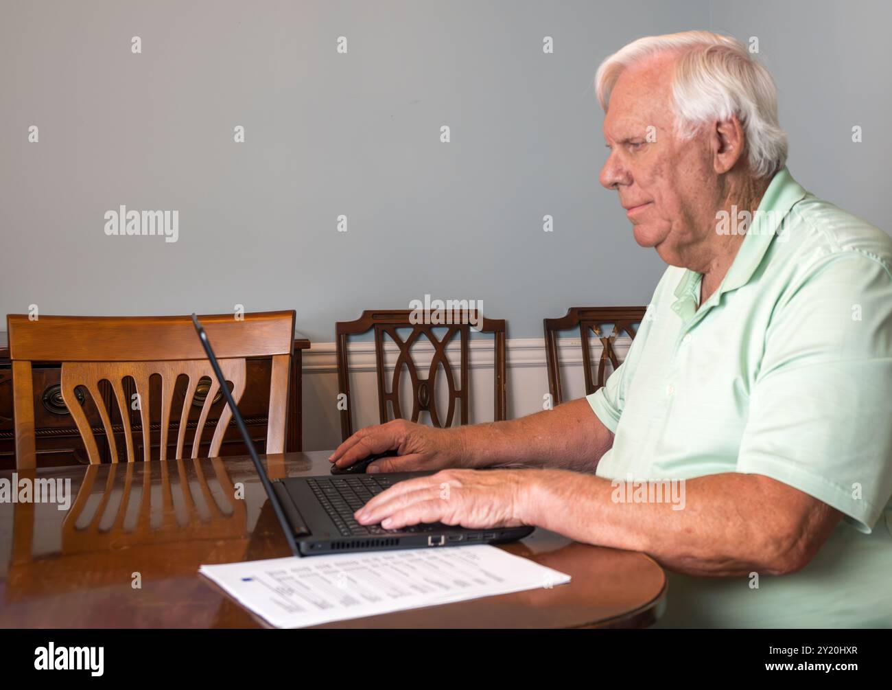 Ein älterer Mann mit weißen Haaren und grünem Hemd arbeitet an seinem Laptop in einem provisorischen Büro in seinem Esszimmer. Stockfoto