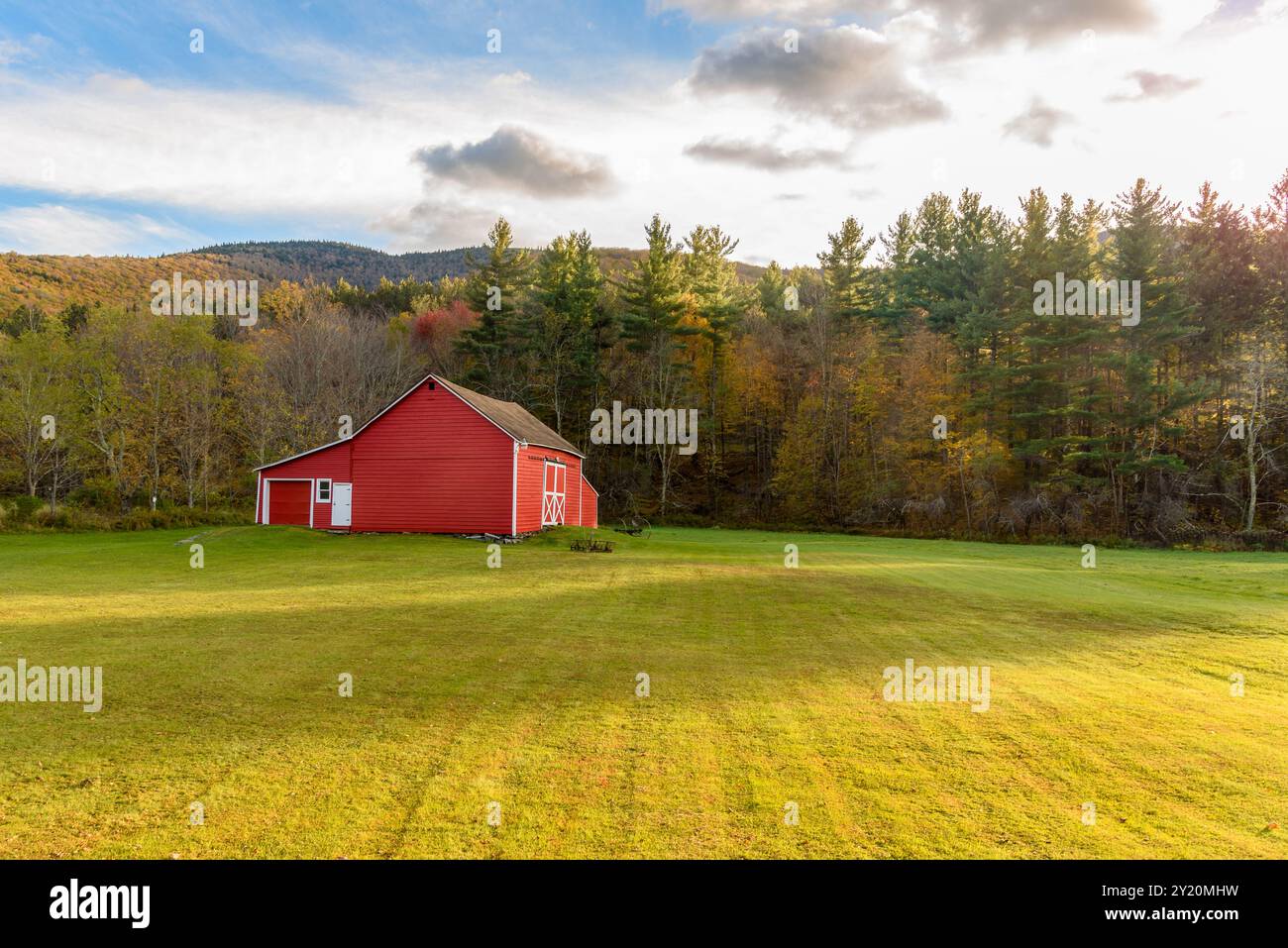 Traditionelle rote Holzscheune am Ende eines grasbewachsenen Feldes, umgeben von farbenfrohen Wäldern in den Bergen bei Sonnenuntergang im Herbst Stockfoto