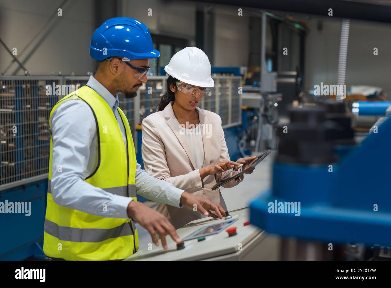 Die indische Betriebsleiterin trainiert einen afroamerikanischen männlichen Mitarbeiter in einem blauen Helm und einer gelben Weste, um Maschinen zu bedienen. Stockfoto