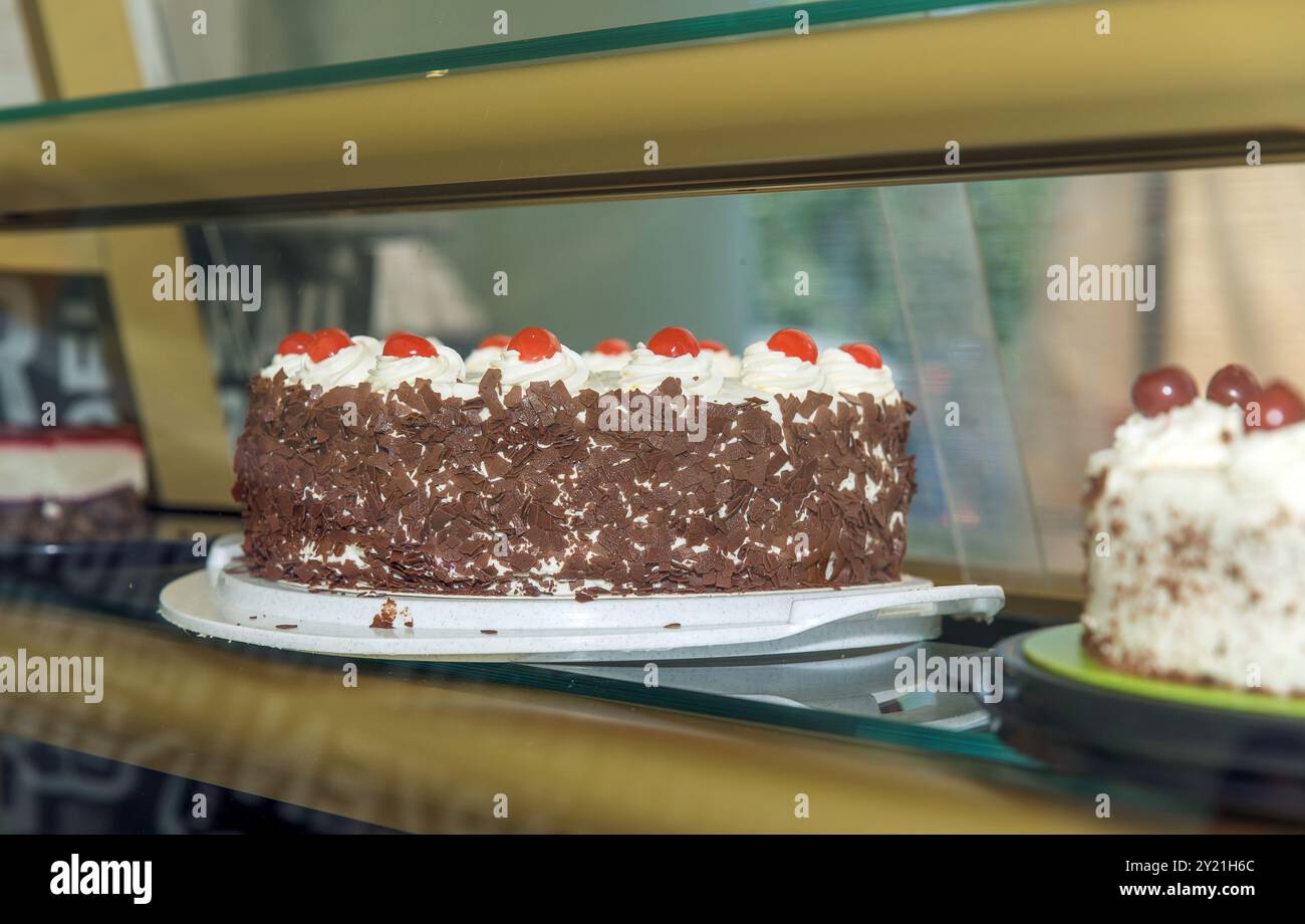 Ein köstlicher Schokoladenkuchen garniert mit Kirschen und cremigem Zuckerguss, ausgestellt auf einem Glasregal in einem Bäckerei-Schaufenster-Konzept Stockfoto