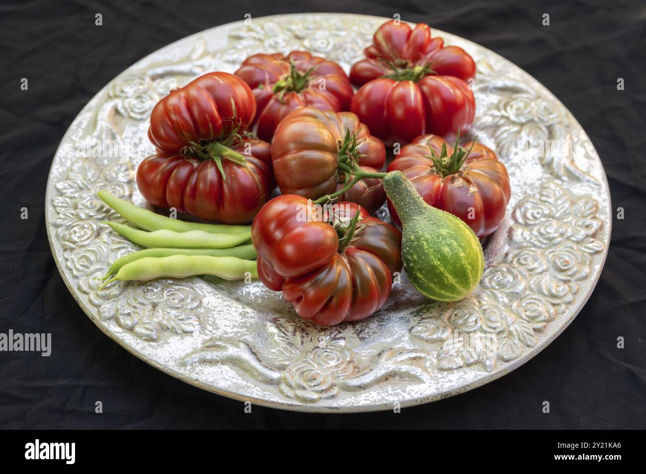 Tomaten, Ochsenherzen und eine in Vorgebirgstrauben eingelegte Gurke auf einem dekorativen Teller auf schwarzem Hintergrund, Baden-Württemberg, Deutschland, Europa Stockfoto
