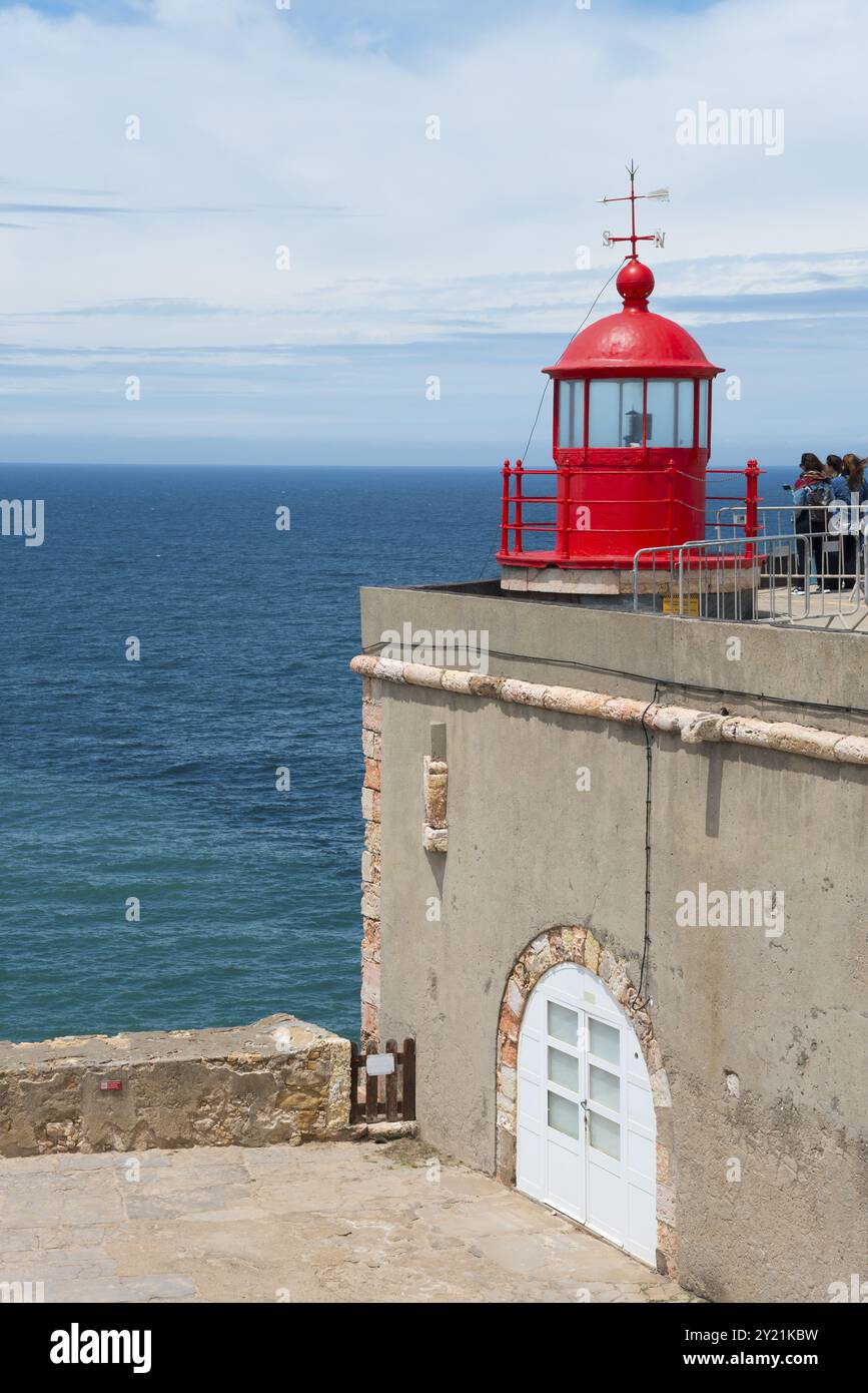 Roter Leuchtturm auf einer Küstenplattform mit Meer im Hintergrund und Menschen, die die Aussicht genießen, Fort Sao Miguel Acanjo, Farol de Nazare, Leuchtturm von Stockfoto