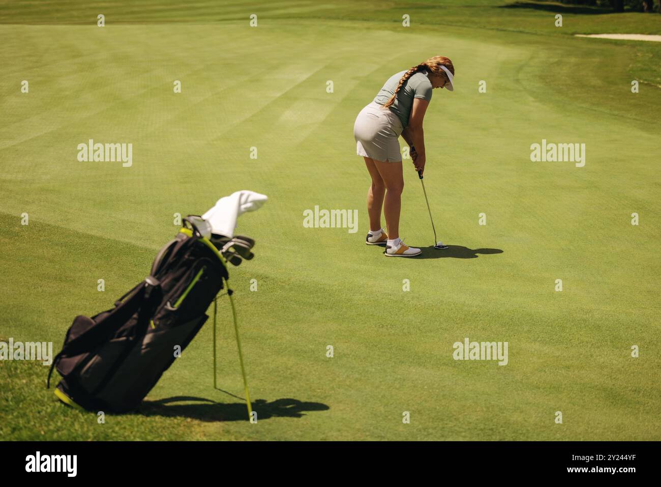 Eine Frau machte sich auf einen Fairway-Schuss vorbereitet, um ihre Haltung und ihren Fokus auf einen sonnigen Golfplatz zu zeigen. Eine Golftasche liegt in der Nähe, während sie sich auf den Ball vorbereitet. Stockfoto