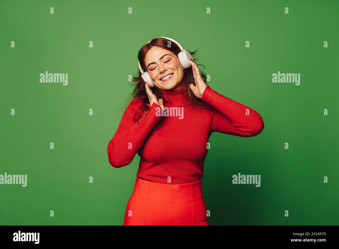 Eine junge Frau in lässiger Kleidung steht selbstbewusst auf einem leuchtenden grünen Hintergrund, trägt ein Noise Cancelling-Headset und hört Musik. Frau enjo Stockfoto