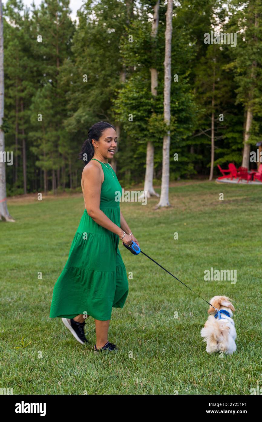 Eine Frau in einem grünen Kleid geht mit ihrem Hund in einem Park. Der Hund ist klein und weiß Stockfoto