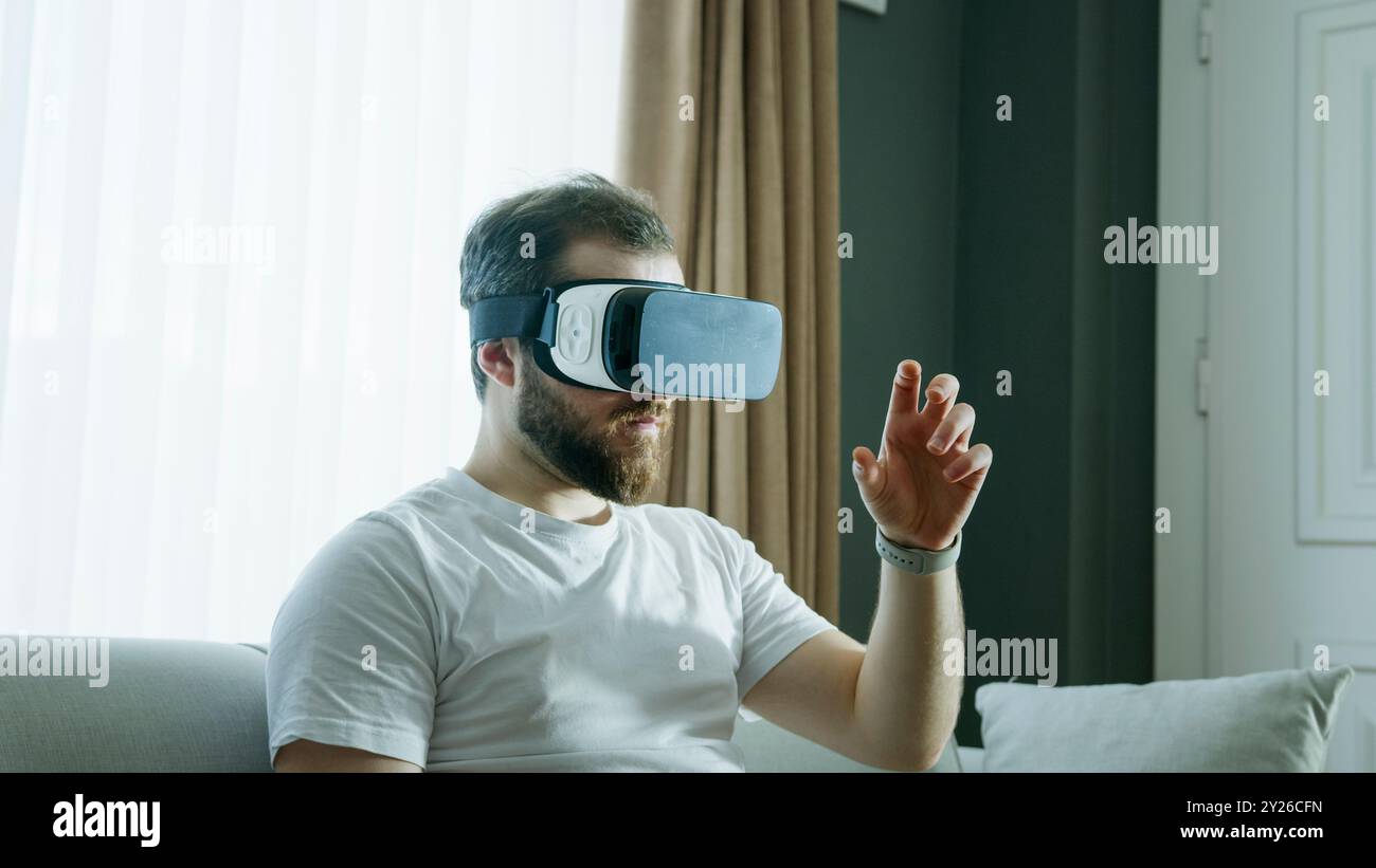 Ein Mann in einem weißen Hemd sitzt auf einer Couch, trägt ein VR-Headset und streckt aus, während er mit der virtuellen Welt interagiert. Die immersive Natur des Schnitts Stockfoto