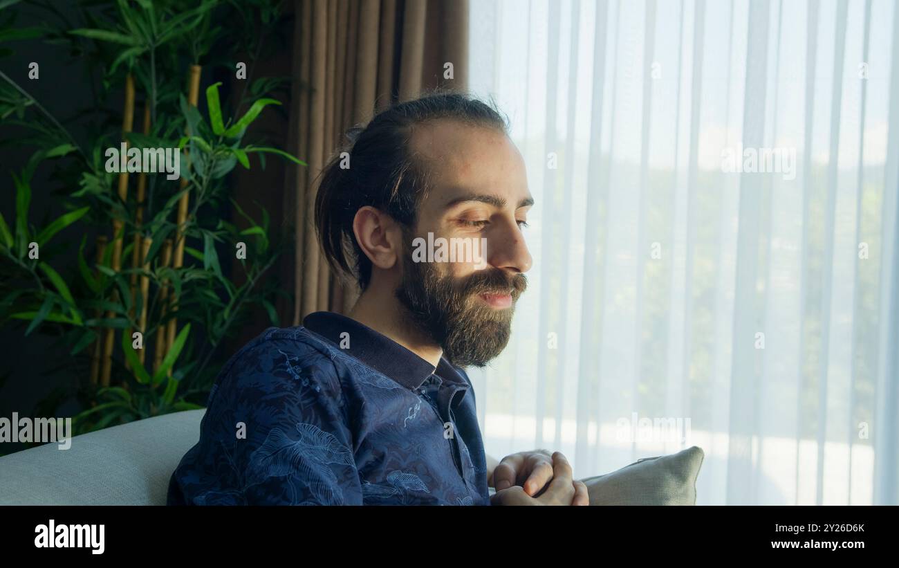 Ein bärtiger Mann sitzt auf einem Sofa und genießt das Sonnenlicht, das durch das Fenster kommt. Ruhige und entspannte, friedliche Atmosphäre in einem gemütlichen Wohnzimmer. Stockfoto