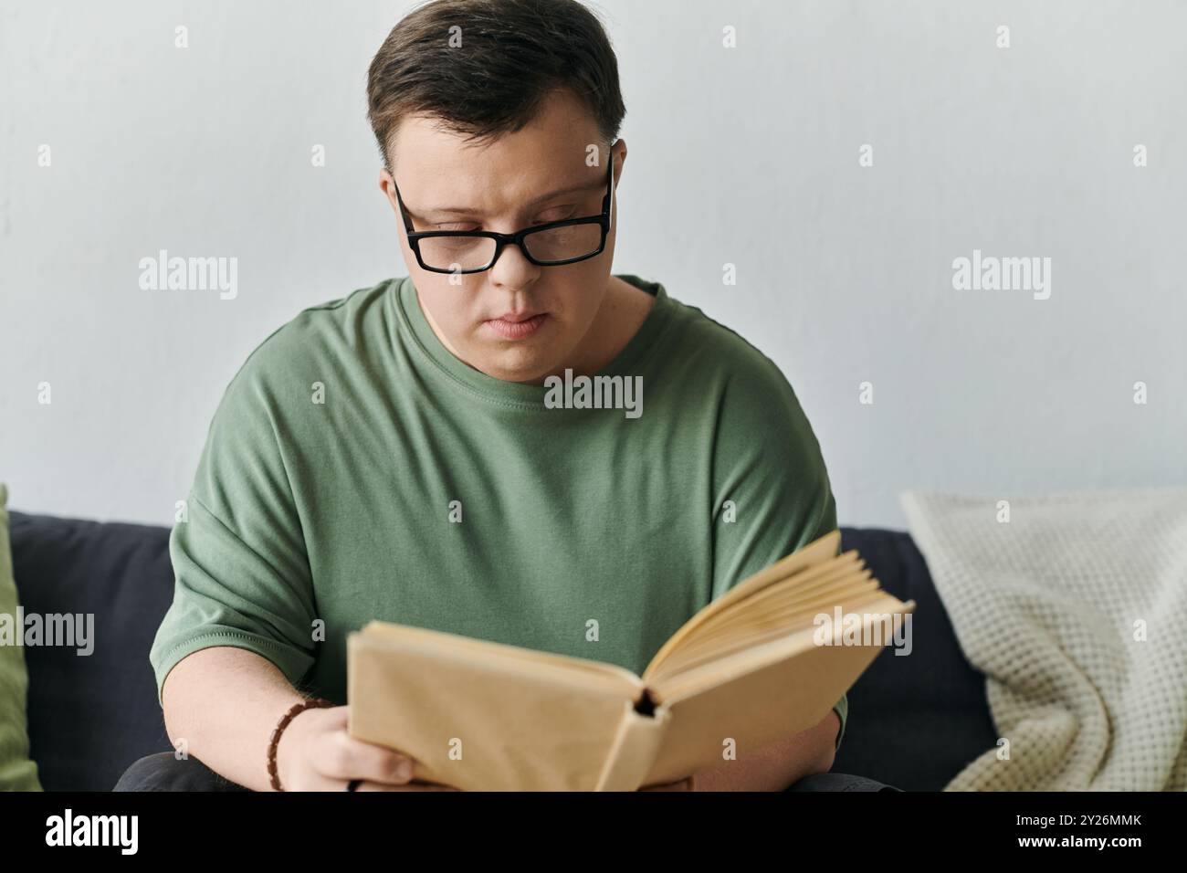 Ein junger Mann mit Down-Syndrom konzentriert sich intensiv auf ein Buch, während er bequem in seinem Wohnzimmer sitzt. Stockfoto