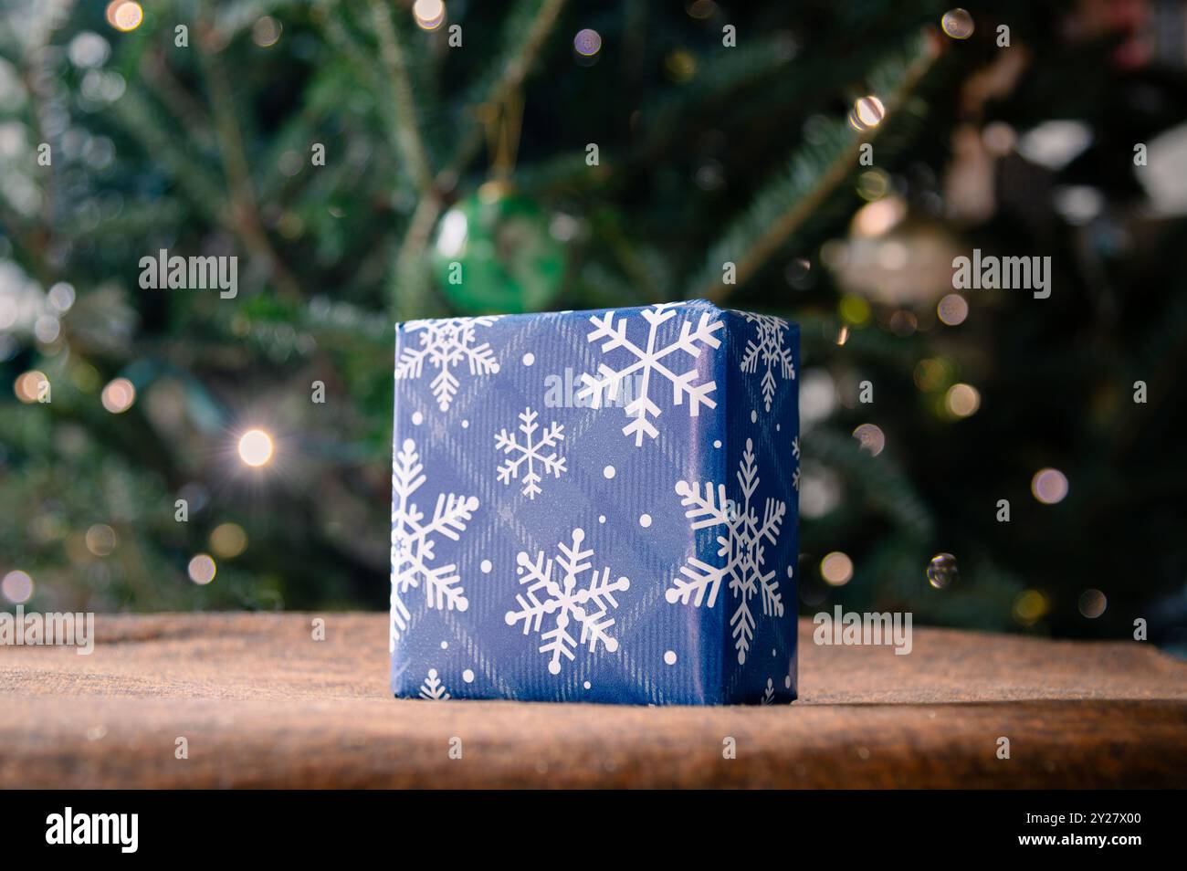 Ein kleines quadratisches Geschenk in blauer Schneeflocke Geschenkpapier mit Weihnachtsbaum im Hintergrund Stockfoto