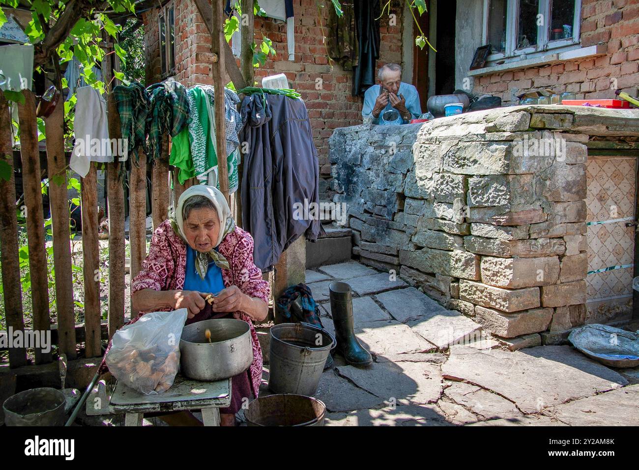 Ein älteres Ehepaar in einem ukrainischen Dorf macht ihre täglichen Aktivitäten. Er rasiert sich, während sie das Abendessen zubereitet. Stockfoto