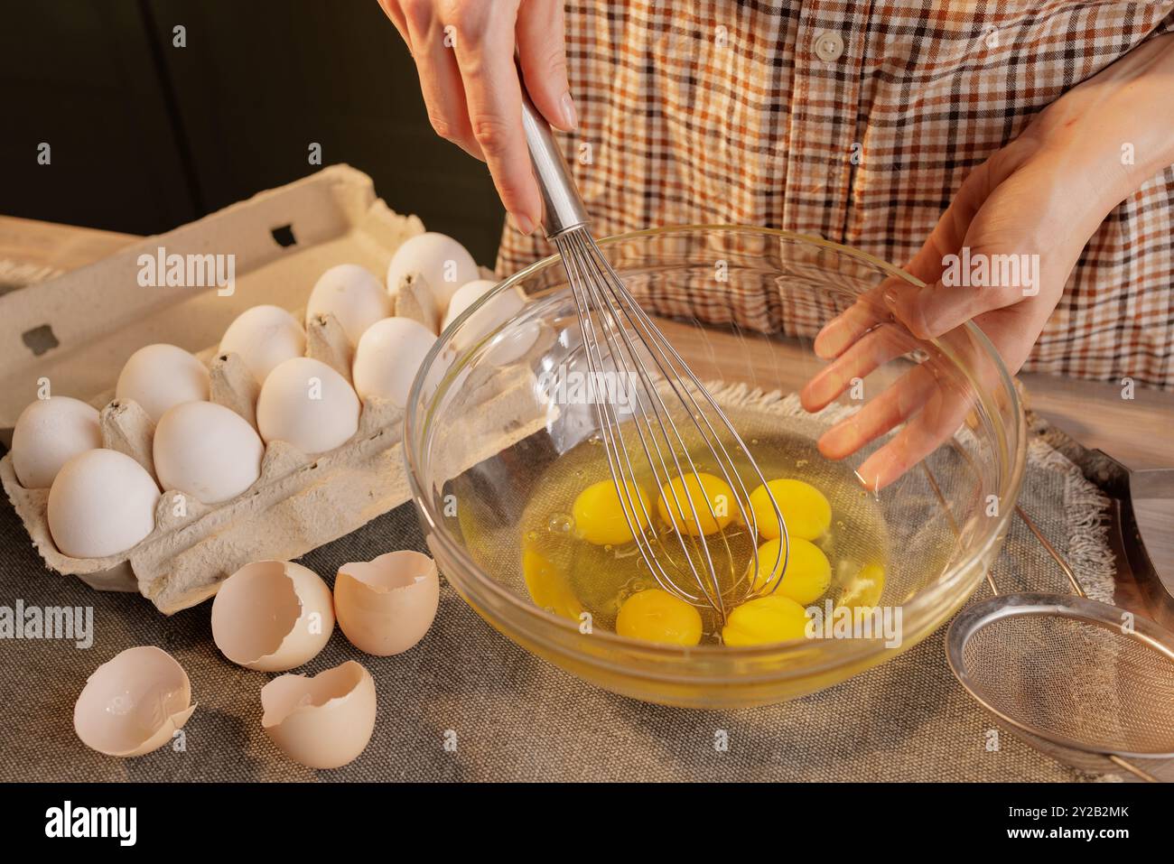 Nahaufnahme von weiblichen Händen, die Eier mit einem Handquirl in einer durchsichtigen Schüssel vor dem Hintergrund eines Küchentischs schlagen. Eier in einer Kiste und ein Werkzeug dafür Stockfoto