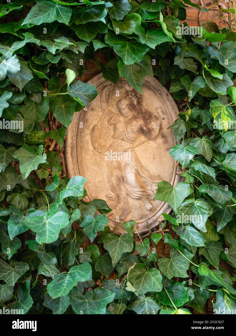Moderne ovale Steinmauertafel, umgeben von Efeu, die eine römische Göttin in einem englischen Garten darstellt. Stockfoto