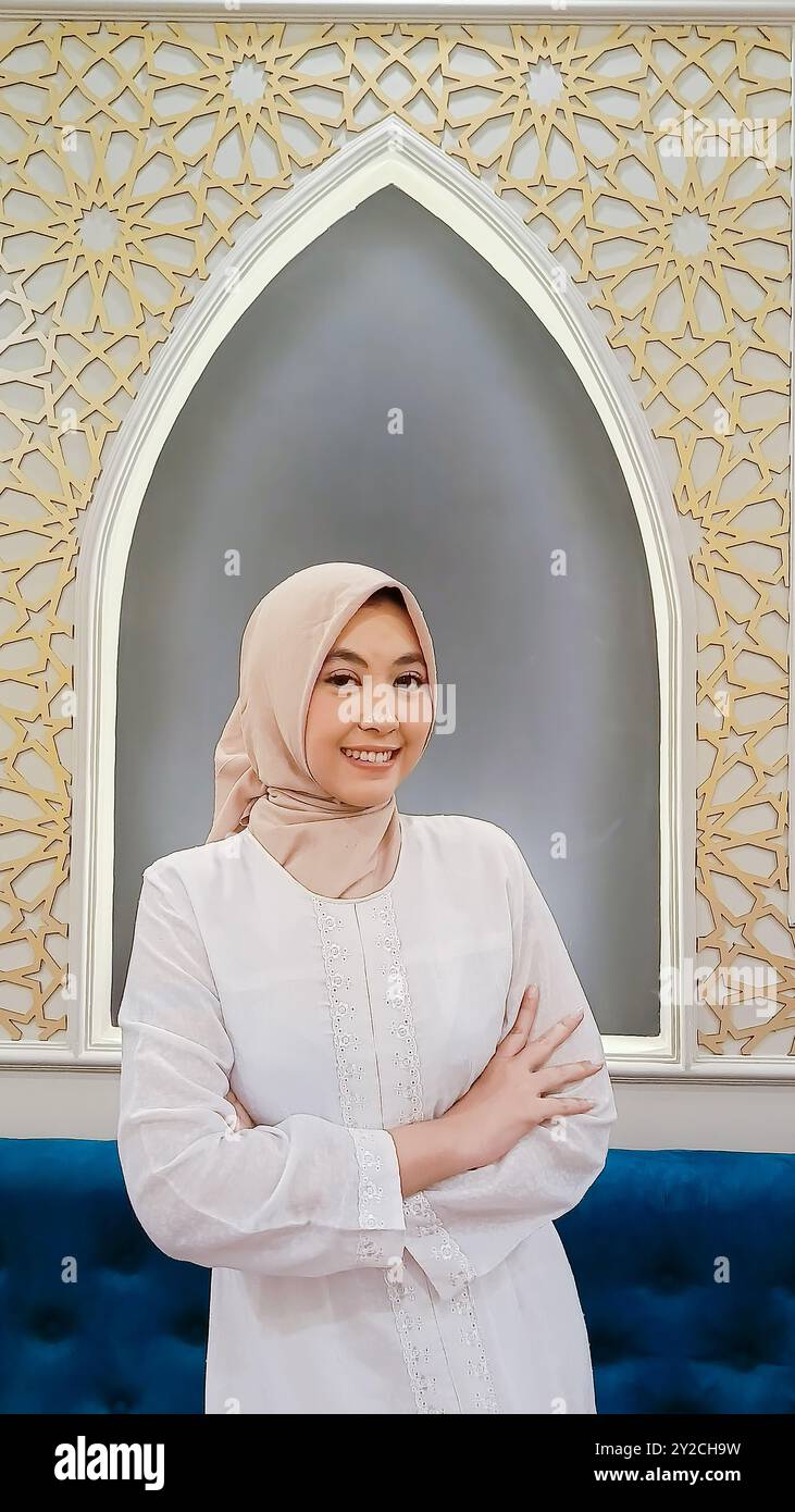 Schönheit der indonesischen Frau, die Hijab trägt, fröhliches Lächeln, Arme mit islamischem Ornament-Kalligraphie-Hintergrund überkreuzt Stockfoto