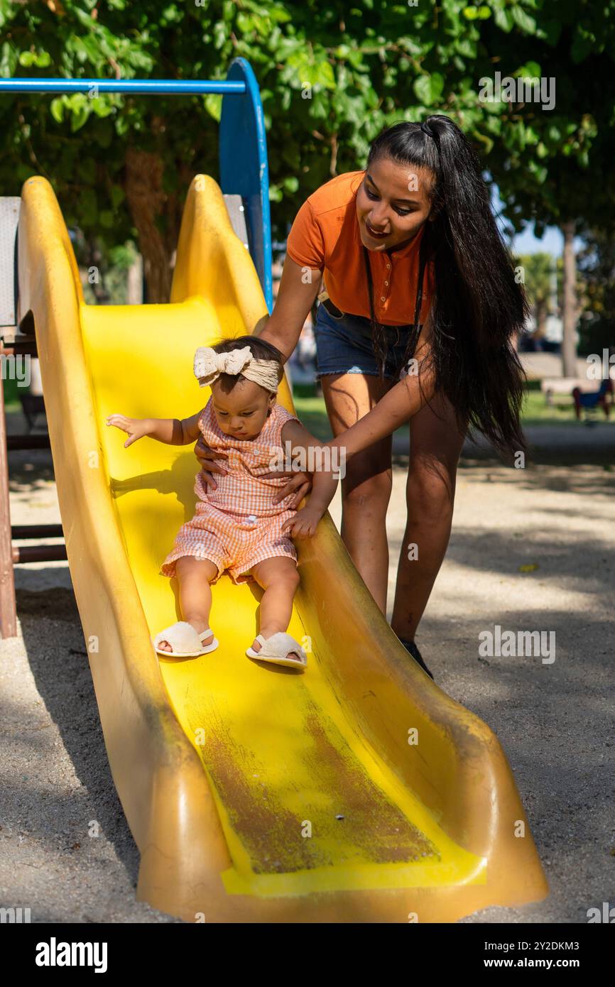 Lateinische Mutter mit ihrer einjährigen Tochter auf der Rutsche eines Spielplatzes Stockfoto