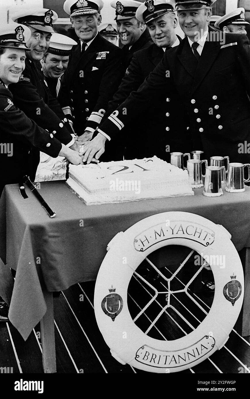 AJAXNETPHOTO. JANUAR 1975. PORTSMOUTH, ENGLAND. - 21. GEBURTSTAG - DIE ROYAL YACHT BRITANNIA IST 21 JAHRE ALT; SIE HAT 30 MAL AUF DER GANZEN WELT GEDAMPFT. AUS DIESEM ANLASS FÜHRTE KONTERADMIRAL RICHARD TROWBRIDGE (4. VON LINKS), FLAG OFFCIER ROYAL YACHTS, DEN VORSITZ ÜBER EINEN SPEZIELL GEBACKENEN KUCHEN, DER VON SECHS MITGLIEDERN DER CREW UNTERSTÜTZT WURDE, DIE SEIT IHRER ZEIT BEI BRITANNIA SIND. SIE WURDEN IM JANUAR 1954 IN AUFTRAG GEGEBEN. NACH DEM KUCHENSCHNITT WURDEN SIE JEWEILS MIT EINEM ZINN-BIERKRUG PRÄSENTIERT. FOTO: JONATHAN EASTLAND/AJAX REF:750103 MX340 241408 30 Stockfoto