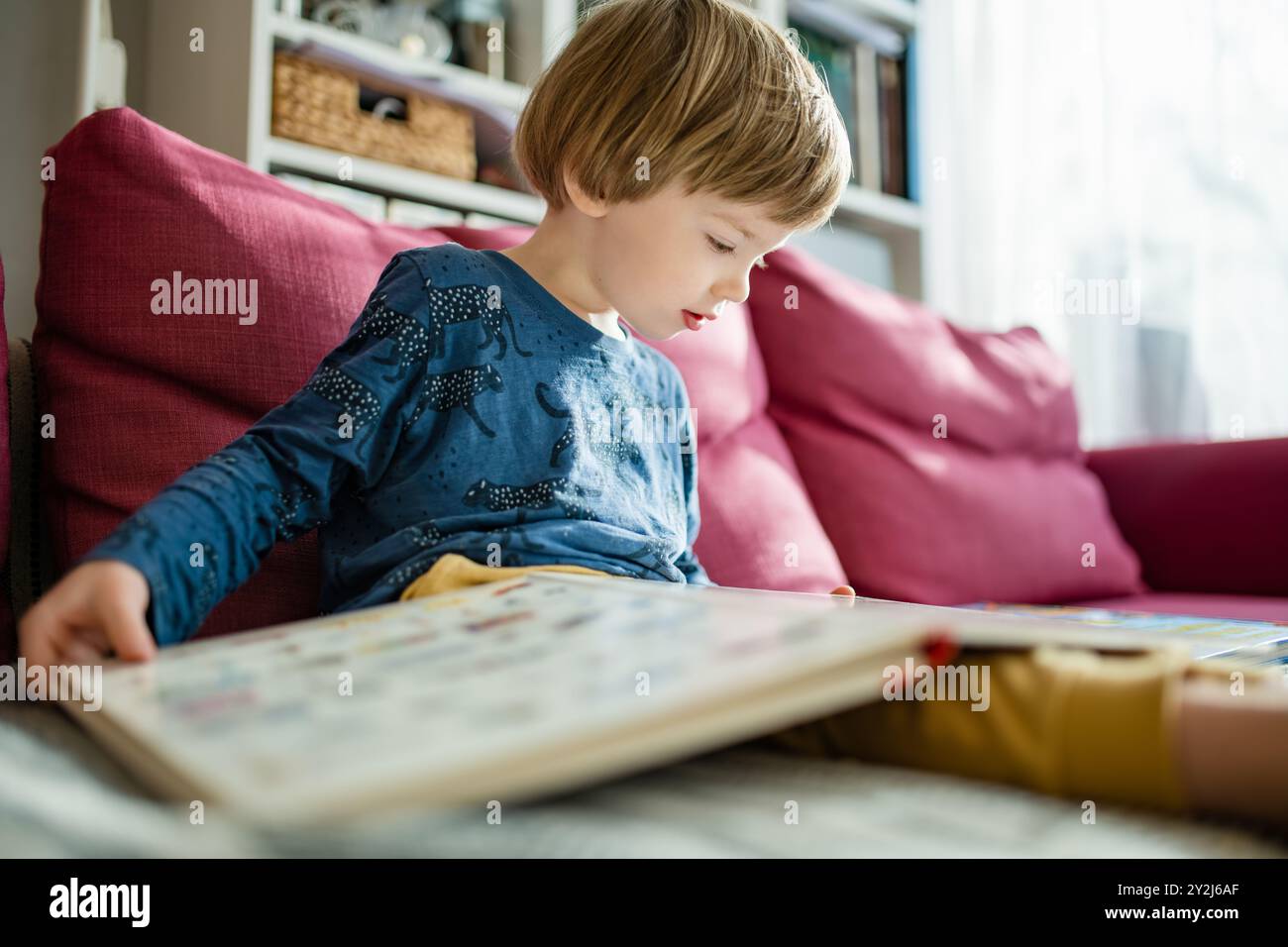 Süßer kleiner Junge, der Bücher auf dem Sofa zu Hause liest, an einem sonnigen Nachmittag. Tagesbetreuung kreative Aktivität. Kinder haben Spaß. Lerninhalte g Stockfoto