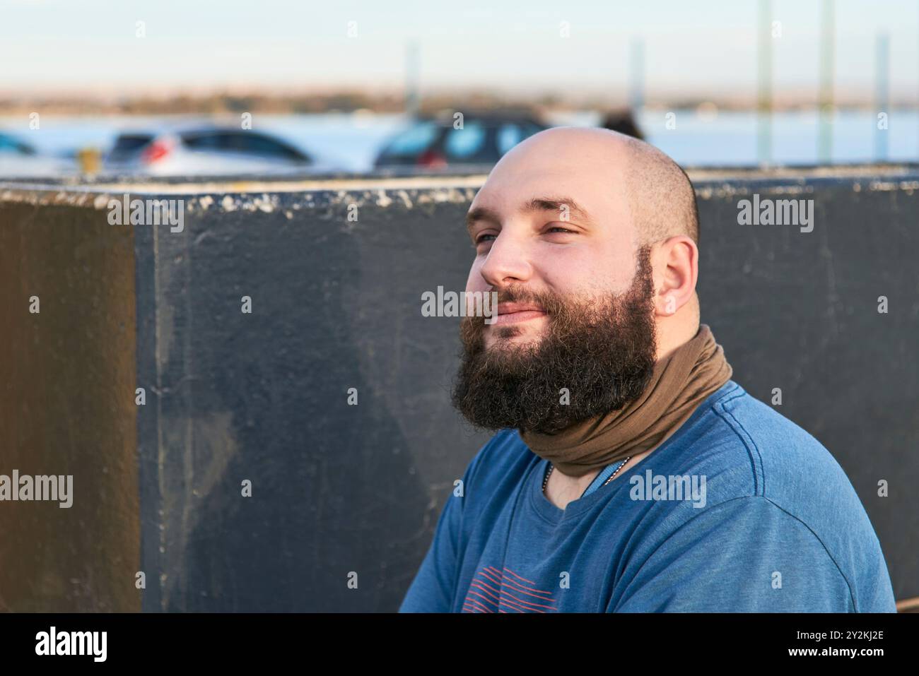 Porträt eines jungen bärtigen Mannes, der draußen sitzt und leicht lächelt, mit einem positiven, ruhigen und freudigen Ausdruck. Komposition mit Kopierraum. Stockfoto