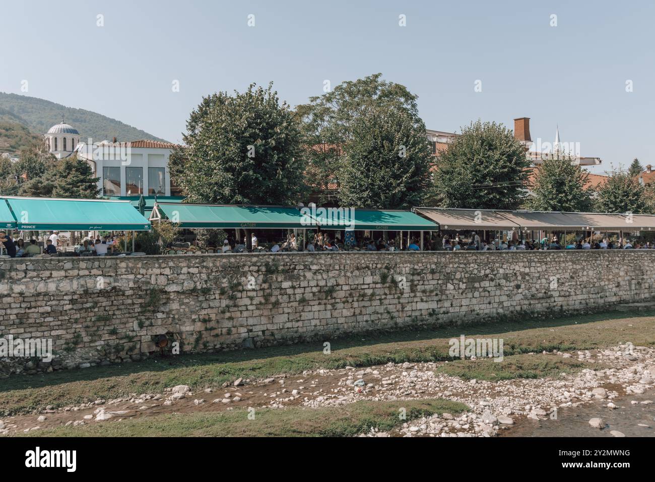 Tische und Stühle vor Bars, Cafés und Restaurants am Fluss Bistrica in der Altstadt von Prizren im Kosovo an einem sonnigen Tag. Stockfoto