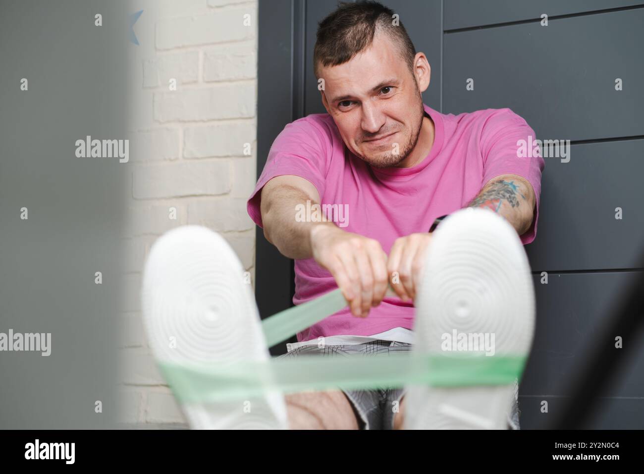 Junger Mann in einem pinkfarbenen Hemd, der zu Hause mit Widerstandsbändern trainiert und sich während des Workouts in der Halle auf Fitness konzentriert. Stockfoto