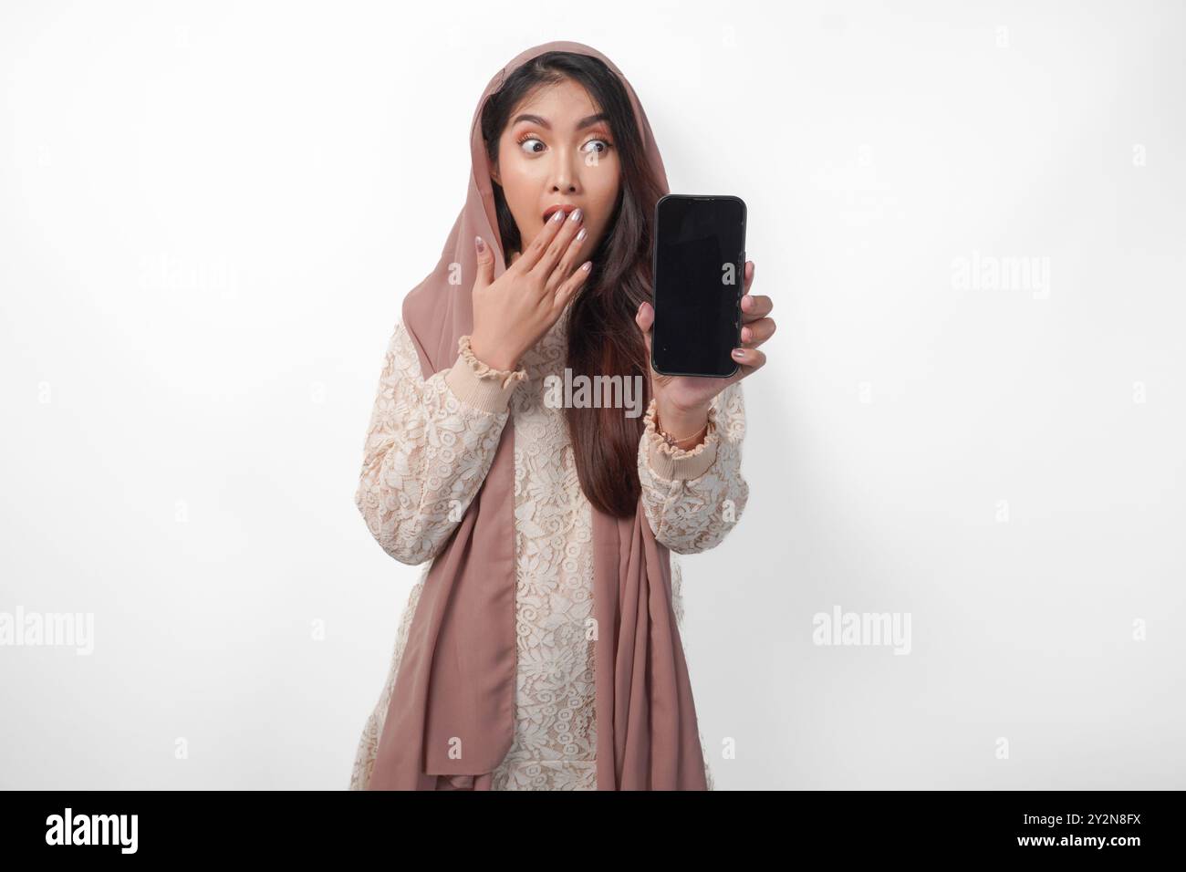 Schockierte asiatische muslimische Frau im Kopftuchschleier-Hijab mit der Hand, die den Mund bedeckt, während sie einen leeren Telefonbildschirm mit einem Kopierraum zeigt, isolierter weißer Hintergrund Stockfoto