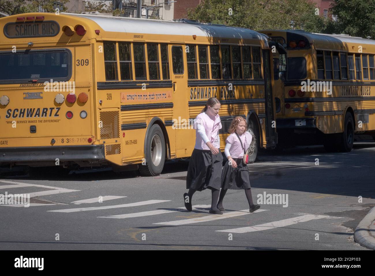 Zwei bescheiden gekleidete orthodoxe jüdische Mädchen überqueren die Straße, vorbei an geparkten Schulbussen in Brooklyn, New York. Stockfoto