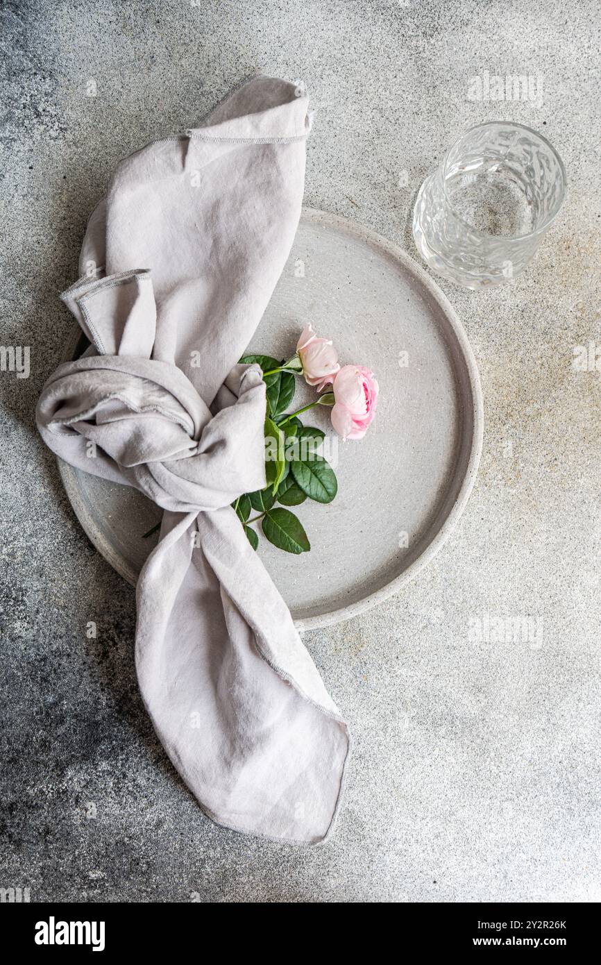 Eine minimalistische Komposition mit zarten rosa Rosen und einem Knoten grauer Leinenserviette, auf einer strukturierten Keramikplatte mit einem klaren Glas im Mais Stockfoto