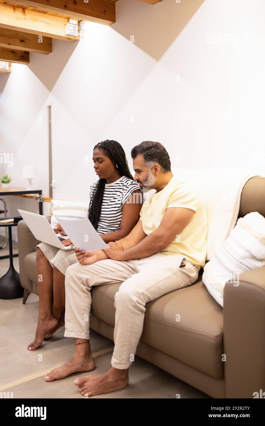 Ein multiethnisches Paar sitzt auf einem Sofa und arbeitet gemeinsam an einem Laptop in einem gemütlichen, gut beleuchteten Wohnzimmer und zeigt eine entspannte und kollaborative Umgebung Stockfoto