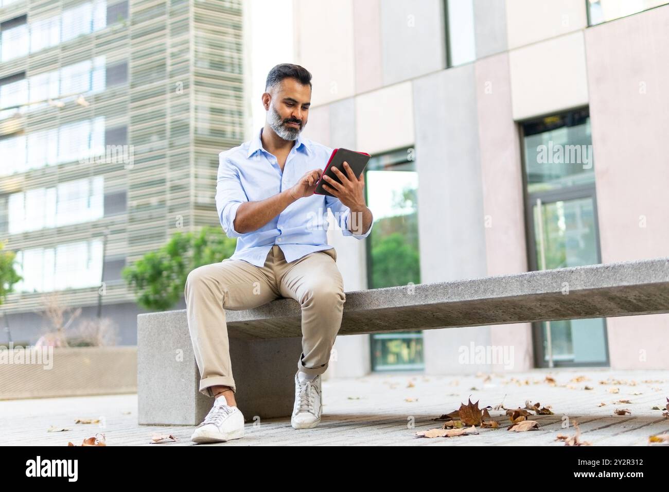 Indischer Geschäftsmann konzentrierte sich auf Tablets, während er auf einer Bank in einer modernen Stadtumgebung saß und in Business-Freizeitkleidung gekleidet war. Stockfoto
