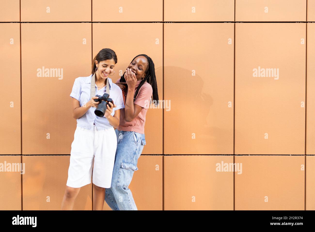 Zwei Collegefreunde, einer hält eine Kamera und freuen sich, wenn sie gemeinsam Fotos vor einem einfachen orangen Hintergrund betrachten und das Wesen des Jugendlichen festhalten Stockfoto