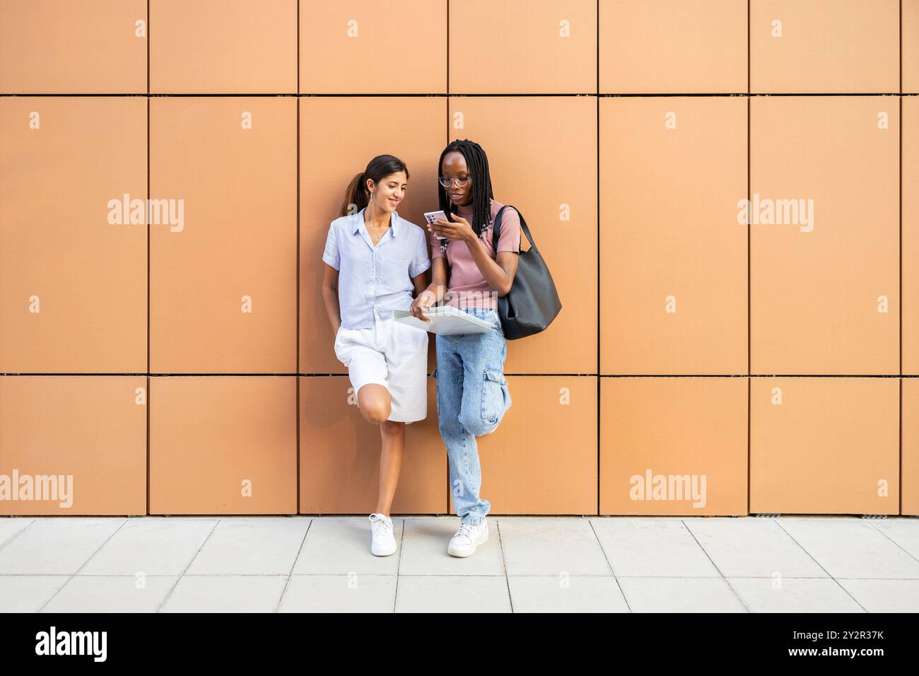 Zwei junge Studentinnen teilen sich einen Moment, während sie ein Smartphone vor einem modernen orangen Hintergrund verwenden. Sie haben einen lässigen Stil und sind in einer Runde aktiv Stockfoto