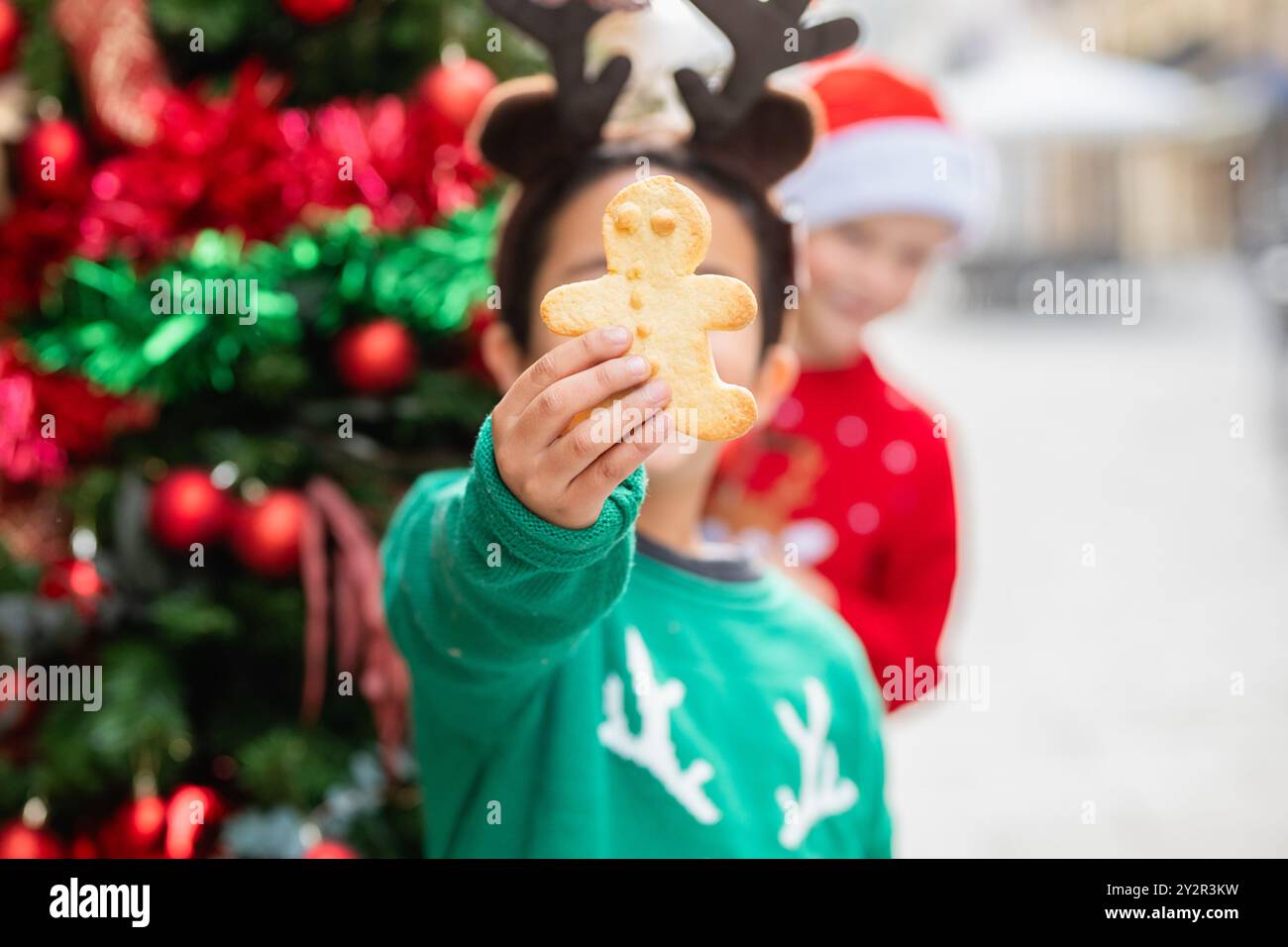 Zwei Kinder in festlicher Kleidung, eines hält einen Lebkuchenkeks vor einem wunderschön geschmückten Weihnachtsbaum Stockfoto