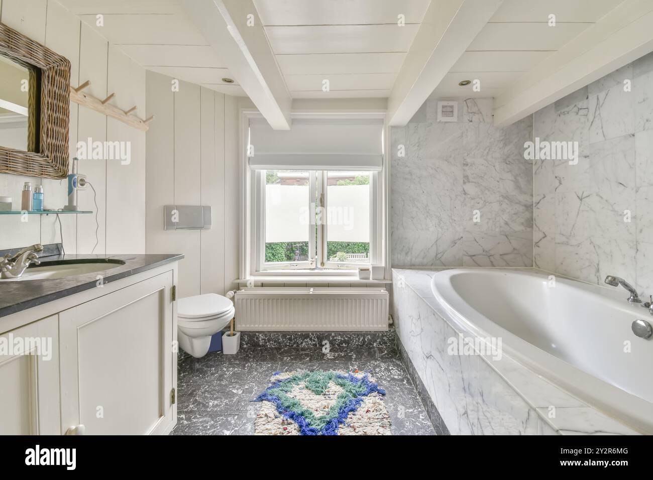 Ein helles und stilvolles Badezimmer mit Marmorfliesen, eingebauter Badewanne, weißen Schränken und einem Fenster mit Gartenblick. Stockfoto