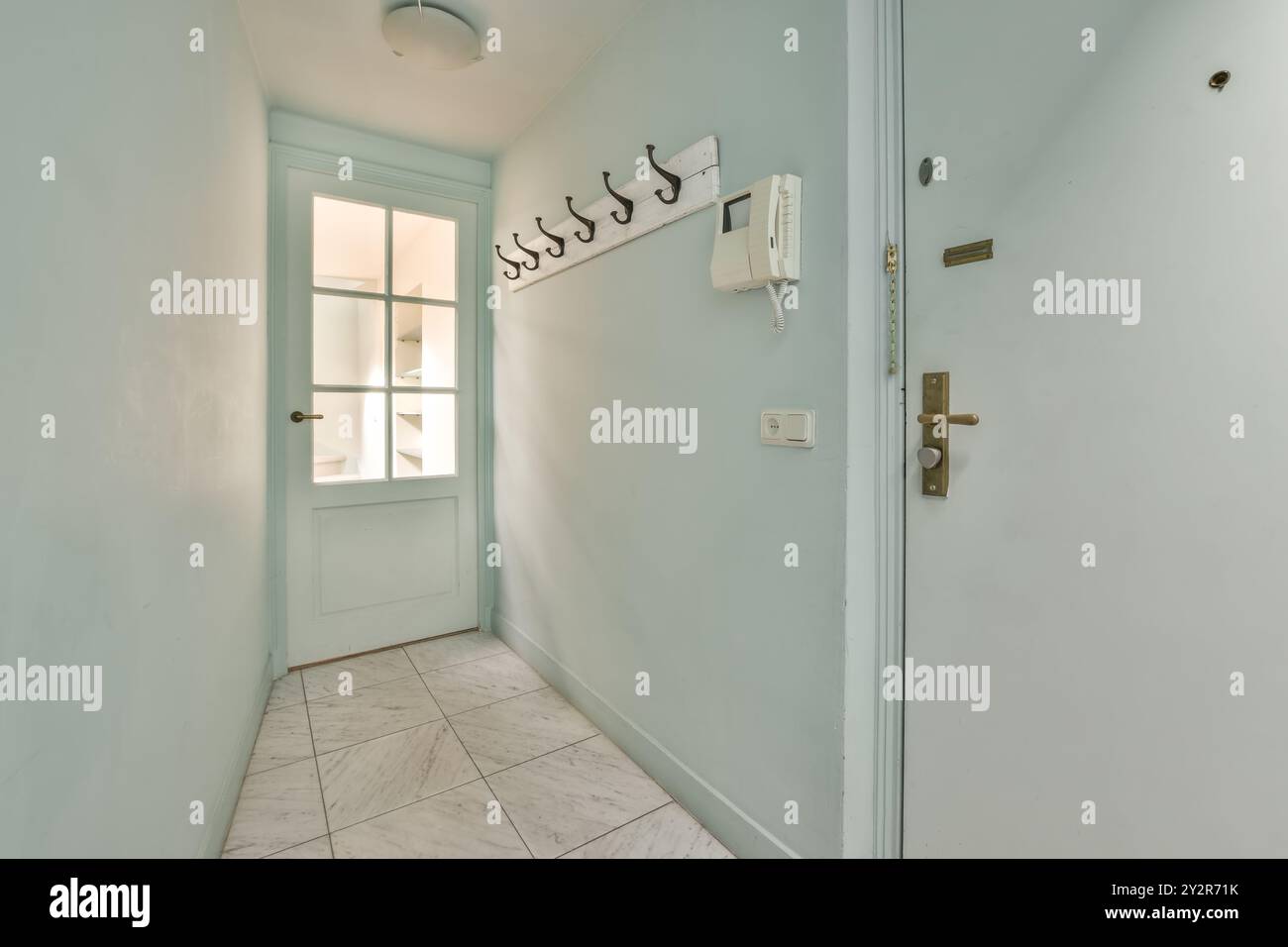 Der minimalistische Flur ist in sanften Blautönen gehalten und verfügt über eine weiße Tür mit Glasfenster, Marmorfußboden und eine Wandgarderobe. Stockfoto