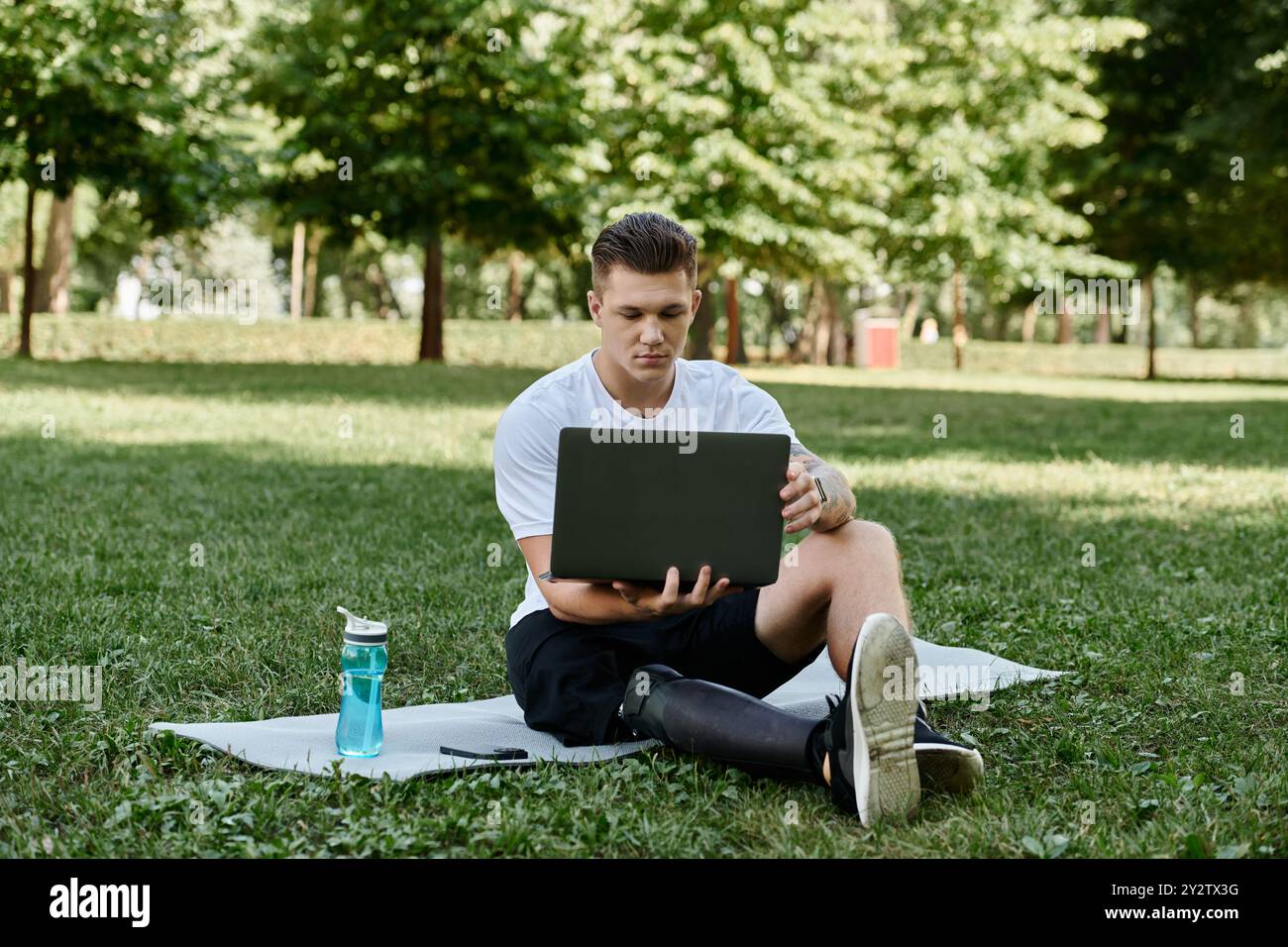 Junger Mann mit Beinprothese und Tätowierungen mit Laptop und auf Fitnessmatte sitzend, digitaler Nomade Stockfoto