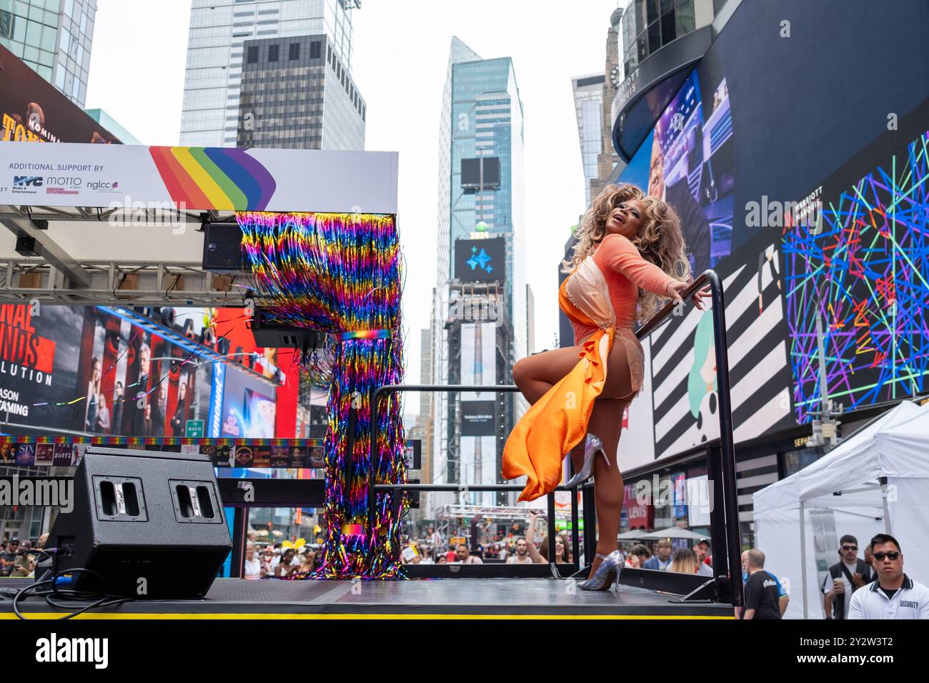 Eine Darstellerin Chicky Gorgina in leuchtendem Orange posiert auf einer Bühne am Times Square, New York City, mit farbenfrohen Dekorationen und Wolkenkratzern Stockfoto